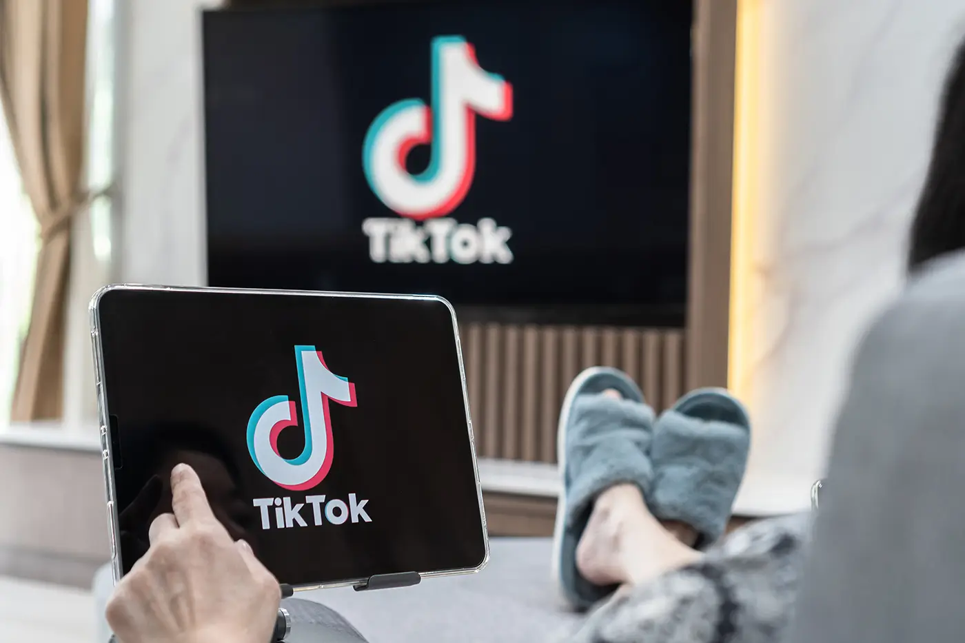 Člověk něco dělá na tabletu s logem sociální sítě a před ním je umístěná televize také s logem stejné sociální sítě