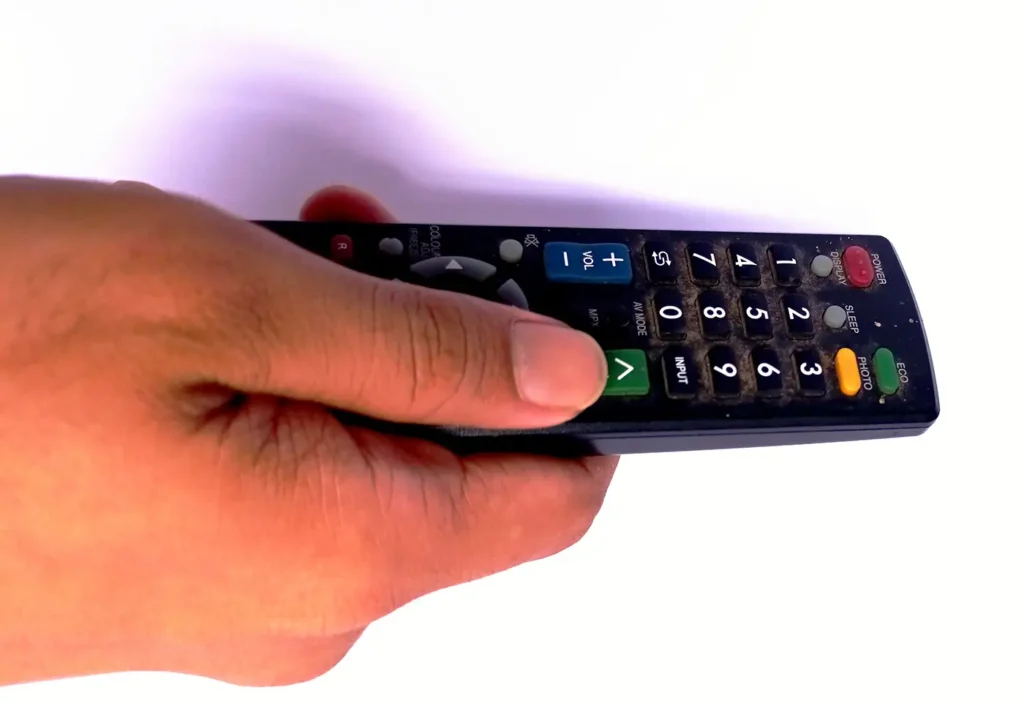 shutterstock_2721316165 - VIPshow Close-up of a hand holding a black TV remote with numeric keypad and colored buttons (green OK, blue volume), angled as if pointing at a screen.