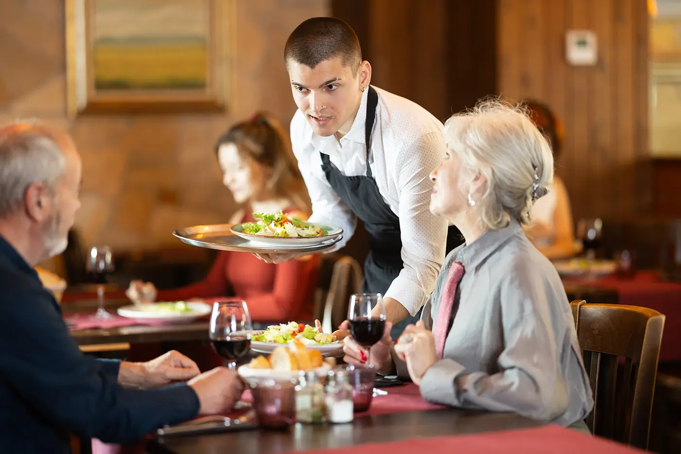 Mladý číšník v restauraci obsluhuje u stolu starší pár