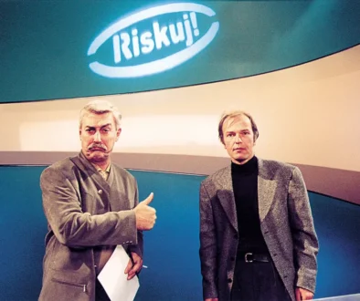 Dva muži stojí ve studiu a nad nimi se skví neonový nápis
