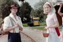 Man in a brown cowboy hat, striped shirt with suspenders and backpack, beside a woman in a pale pink vintage dress with veil, near railroad tracks.