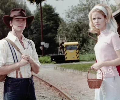 Man in a brown cowboy hat, striped shirt with suspenders and backpack, beside a woman in a pale pink vintage dress with veil, near railroad tracks.