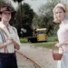 Man in a brown cowboy hat, striped shirt with suspenders and backpack, beside a woman in a pale pink vintage dress with veil, near railroad tracks.