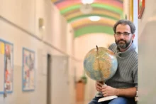 Man with glasses and a gray sweater sits in a hallway, holding a globe and a pointer.