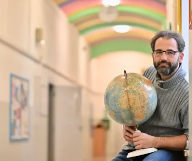 Man with glasses and a gray sweater sits in a hallway, holding a globe and a pointer.