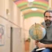 Man with glasses and a gray sweater sits in a hallway, holding a globe and a pointer.