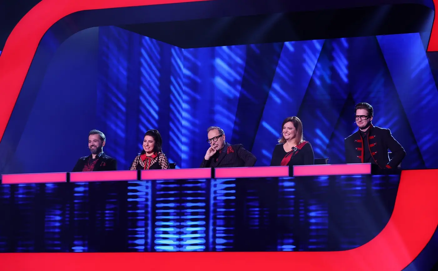 Five judges seated behind pink-lit desks on a blue-lit game show stage, smiling at the camera.",