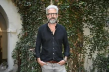 Man with gray hair and beard in a black button-down shirt, standing outdoors in front of an ivy-covered wall.