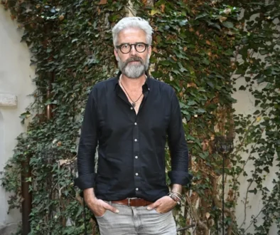 Man with gray hair and beard in a black button-down shirt, standing outdoors in front of an ivy-covered wall.