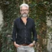 Man with gray hair and beard in a black button-down shirt, standing outdoors in front of an ivy-covered wall.