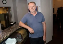 Muž v šedém tričku s límečkem stojí rukou opřený o pult a dívá se před sebe