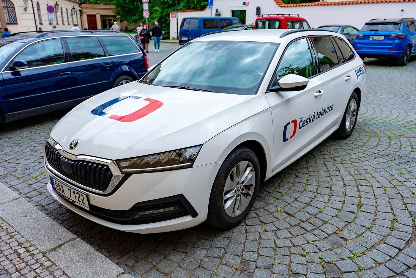 Bílé auto s logem veřejnoprávní televize stojí venku na ulici, na místě určeném pro parkování vozidel