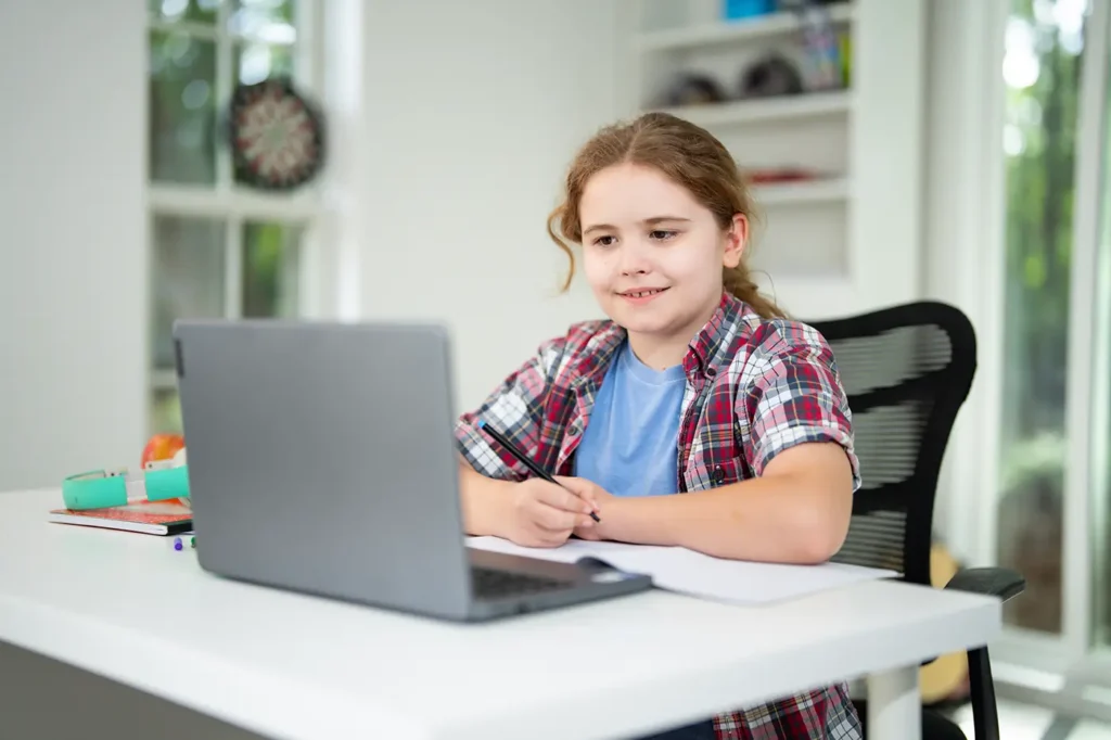 Malá holčička v tričku a košili sedí za stolem a před ní je notebook