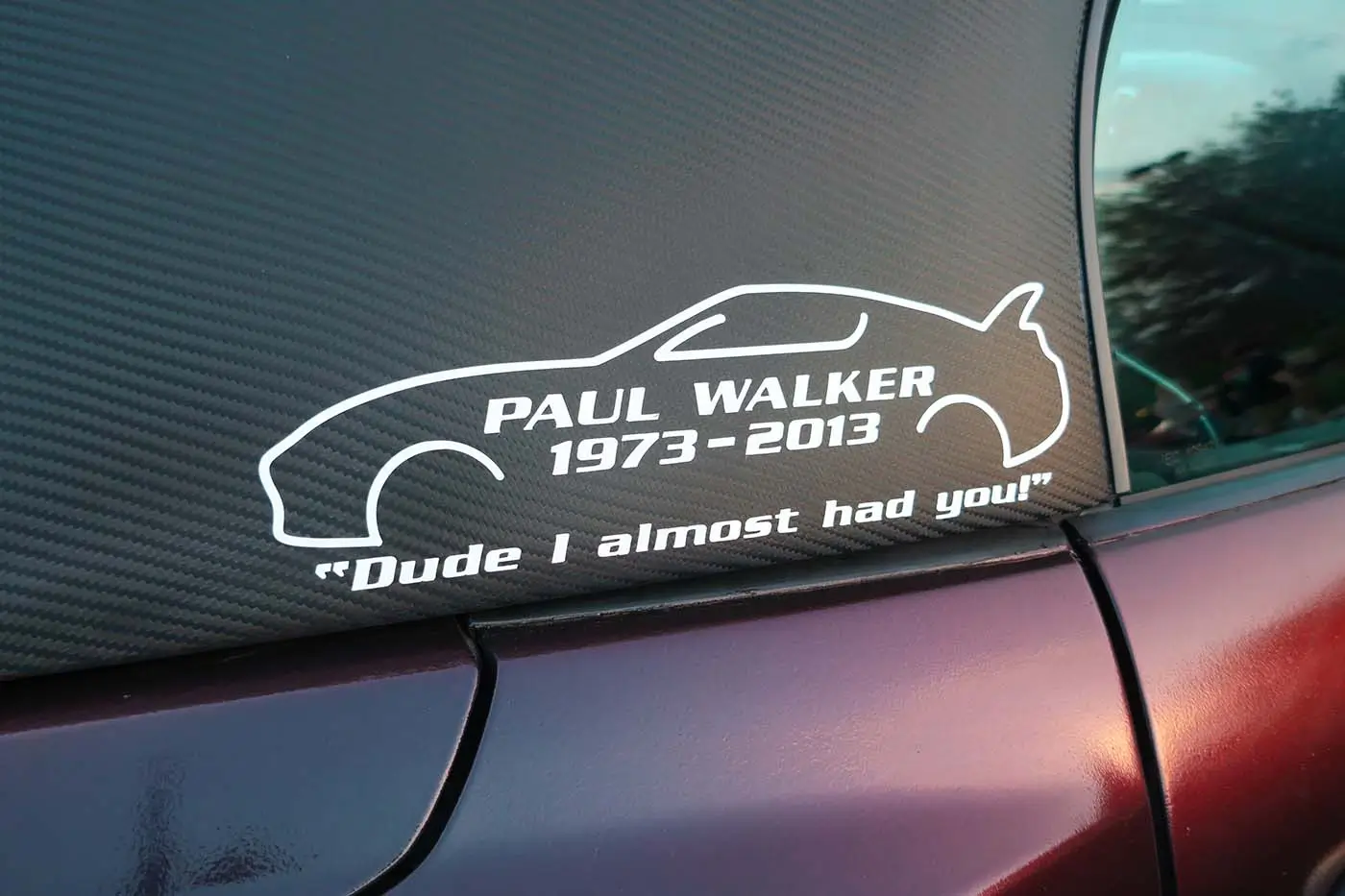 Sticker on a car window showing a stylized car outline with text:'PAUL WALKER 1973-2013' and the quote 'Dude I almost had you!'